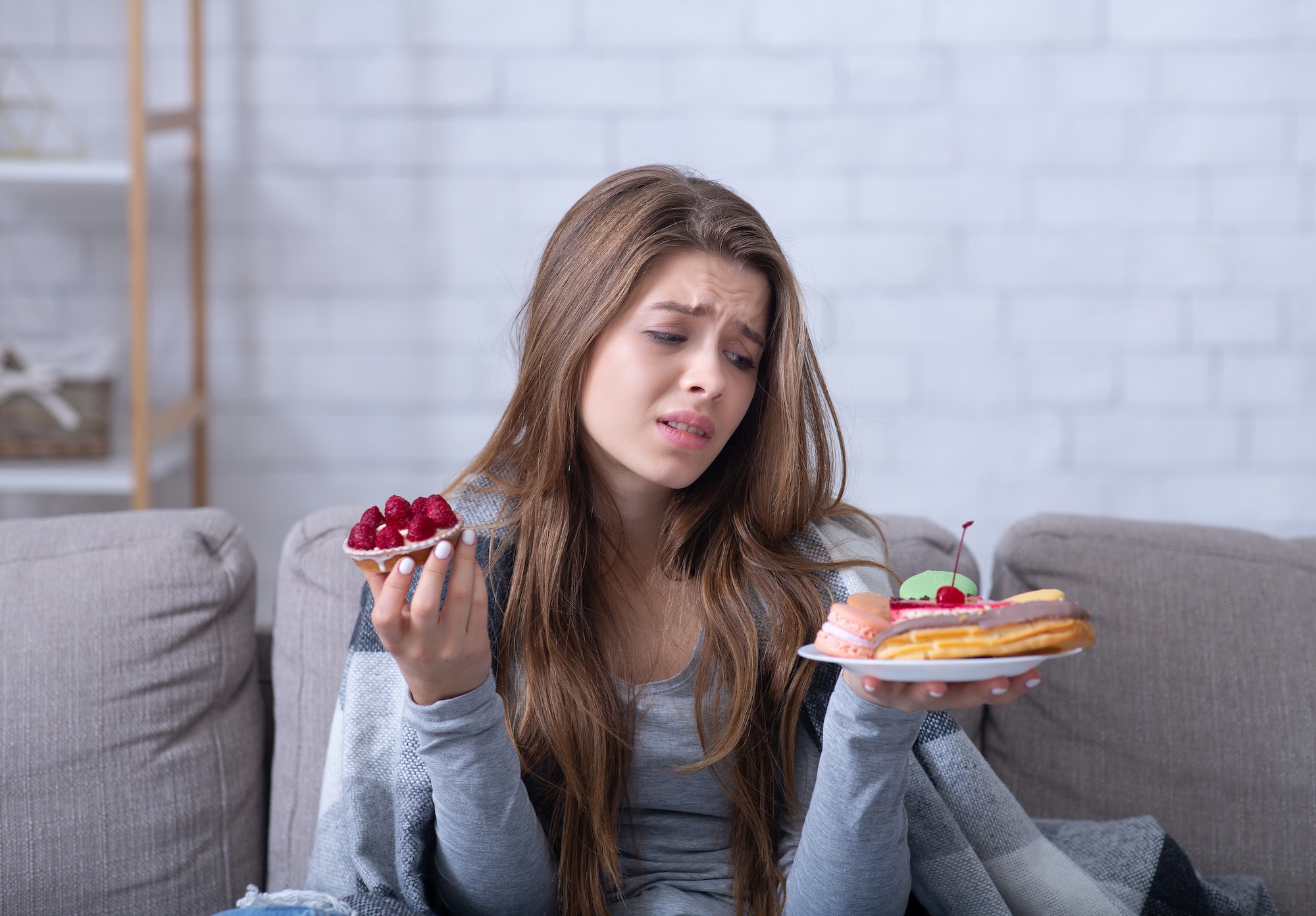 Depressed young lady having eating disorder, comforting herself with sweets on sofa at home